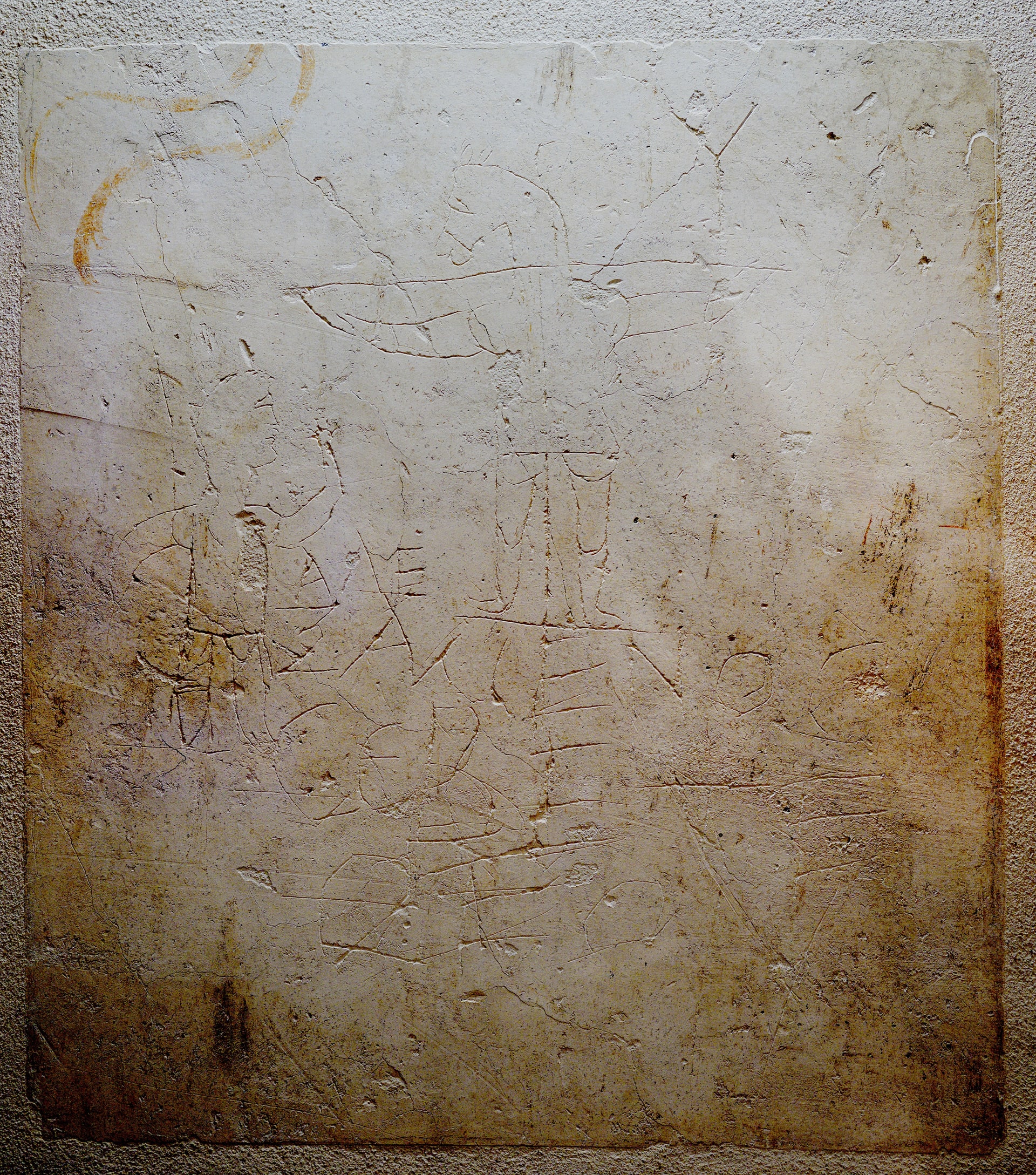 A scratched drawing on plaster shows a man raising an arm toward a crucified, donkey-headed figure, with a Greek inscription below.