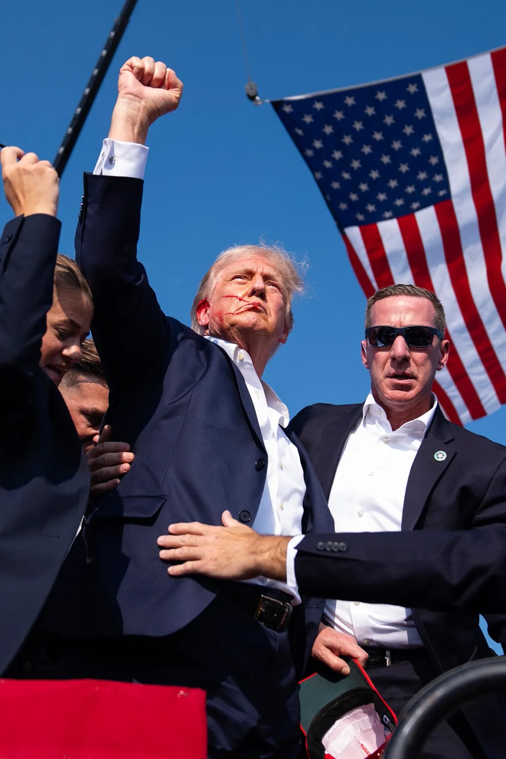 Donald Trump raises a fist among security personnel with an American flag behind them.